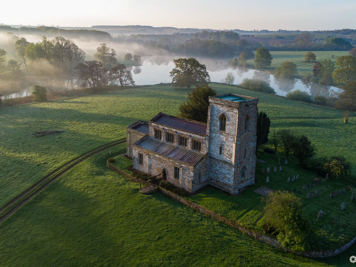 Aerial view of a rural church beside a misty lake at dawn